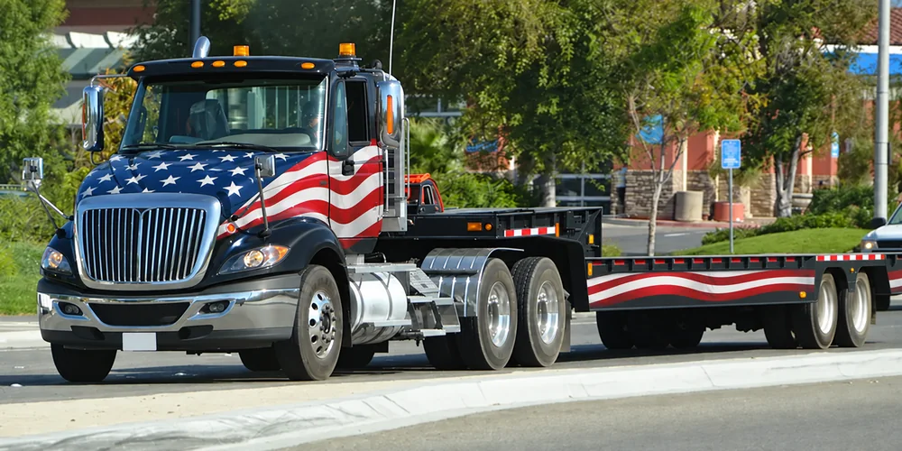 Black equipment-hauling truck with an American flag wrap and lowboy trailer